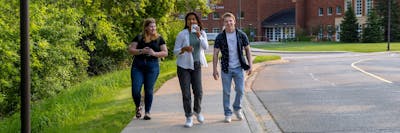  Three college students walking together on a tree-lined campus path, smiling and holding drinks.