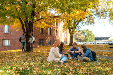 Students studying under fall trees on campus near a brick dorm, with two others talking nearby.