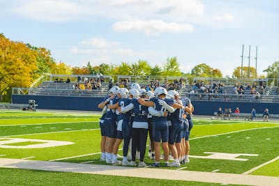 A college football team in navy blue uniforms huddles together on the field before a game, with empty bleachers and autumn-colored trees in the background.