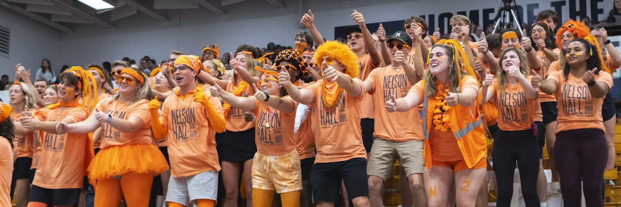 Students in orange Nelson Hall shirts cheer loudly in a packed gym.