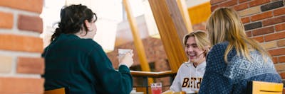 Three students chat and laugh while eating lunch in a sunny campus dining space.
