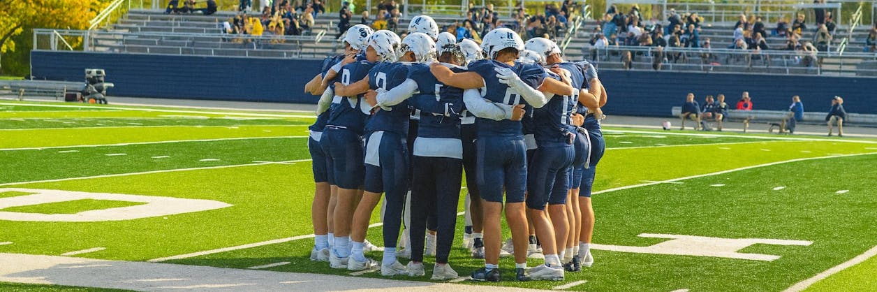 Bethel football players huddle on the field before a game under sunny skies.