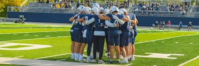 Bethel football players huddle on the field before a game under sunny skies.