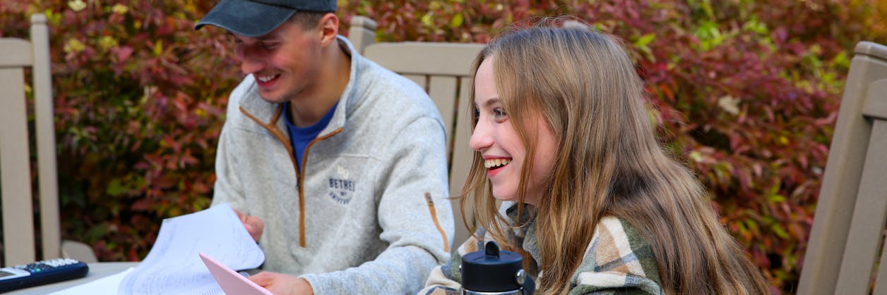 Two students study together outside at a table surrounded by vibrant autumn foliage on Bethel’s campus.