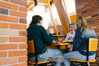 Three Bethel University students share a meal and laughter in the dining hall, surrounded by natural light and warm wooden architecture.