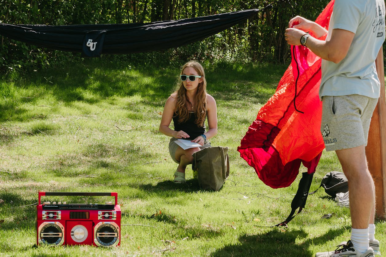 A student sits on the grass with papers and a bag while others set up a hammock nearby on a sunny day.