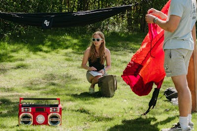 A student sits on the grass with papers and a bag while others set up a hammock nearby on a sunny day.