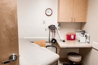 A clinical exam room with a patient bed, medical equipment on the wall, a counter with supplies, and wooden cabinets above.