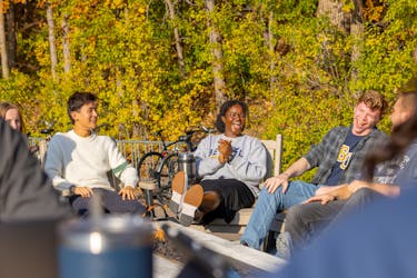 Students laugh and relax around a firepit surrounded by autumn trees at Bethel.