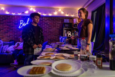 Students enjoy a late-night waffle and pancake event in a cozy dorm lounge with string lights and a neon “Boden” sign, creating a warm and welcoming community atmosphere.