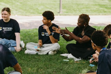 A group of students and a facilitator talk and listen while seated on the grass outdoors at Bethel.