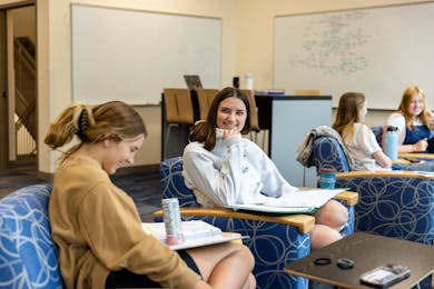 Students smile and study in a bright classroom with notebooks and whiteboards.
