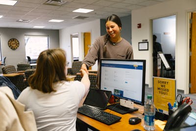 A smiling student shakes hands with someone behind a front desk in a welcoming campus office.
