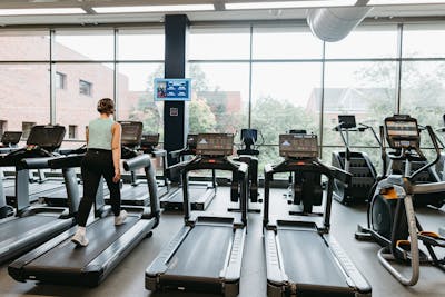 Student walks on a treadmill facing large windows overlooking Bethel’s brick campus buildings.